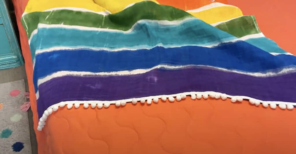 Rainbow blanket tutorial – Learn just how easy it is to make this cute and colorful decorative throw with festive pom pons.