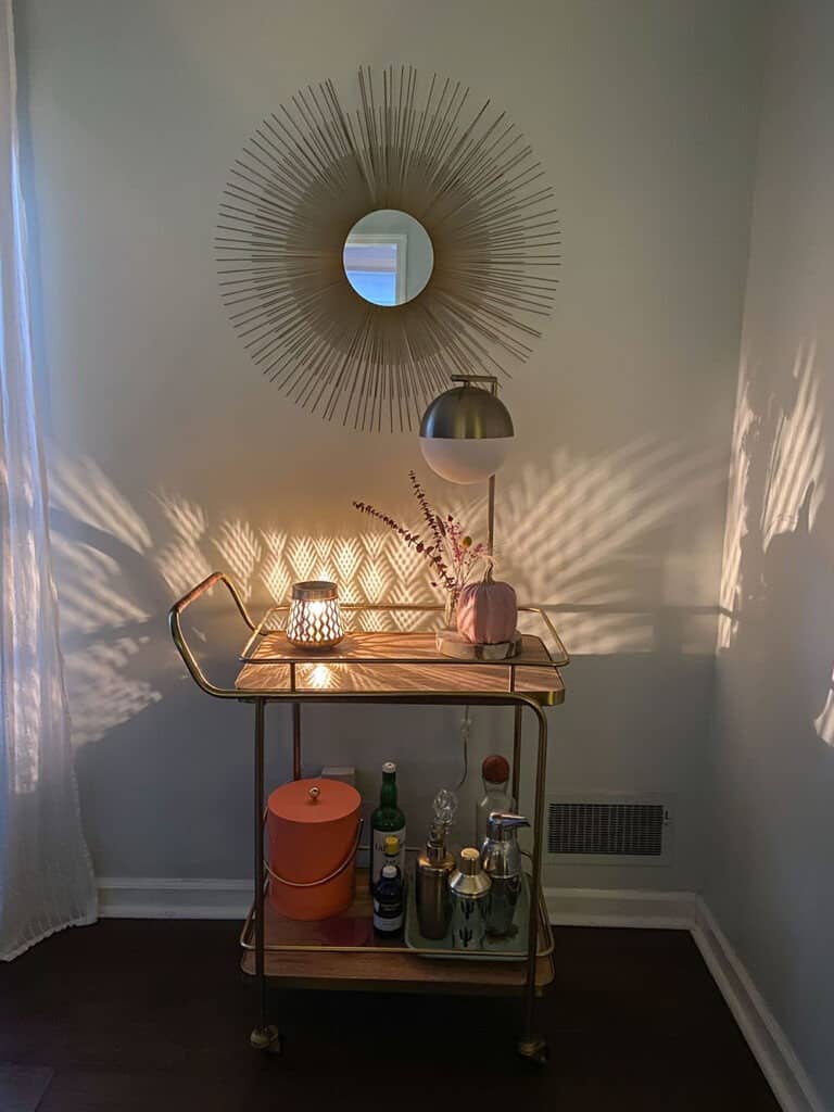 Gold and wood decorated bar cart with a faux pink pumpkin, dried florals in a vase, and a wax warmer that scatters light across the wall