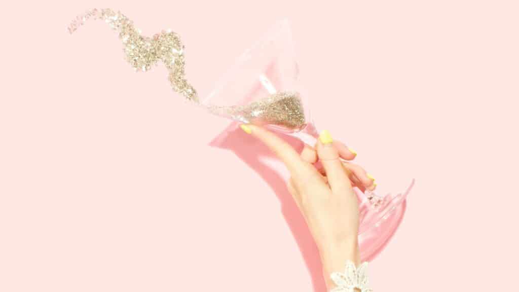 hand holding martini of gold glitter spilling out on light pink background